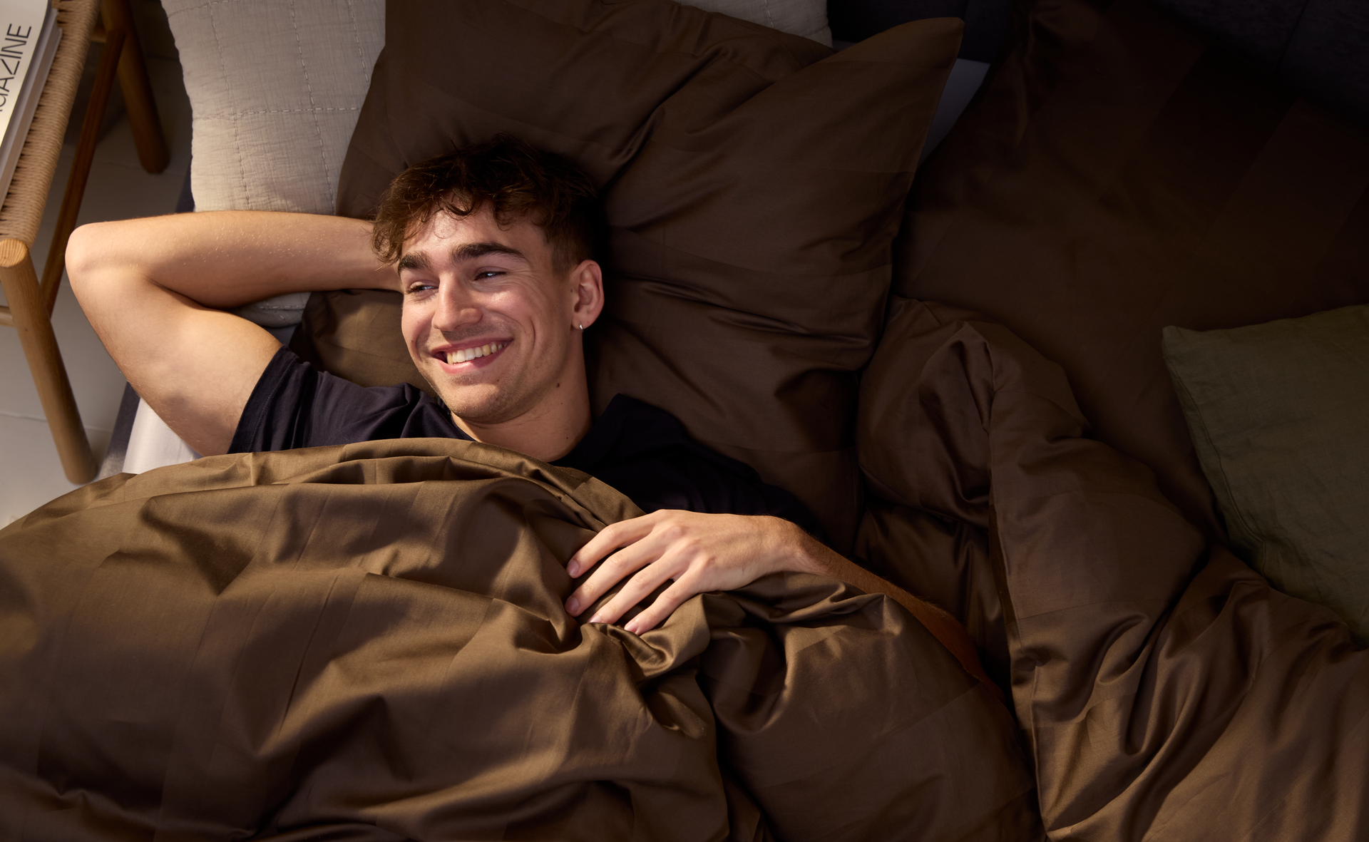 Ein Mann liegt auf einem Bett mit brauner Bettwäsche, den Kopf auf den Arm gestützt, und lächelt. 