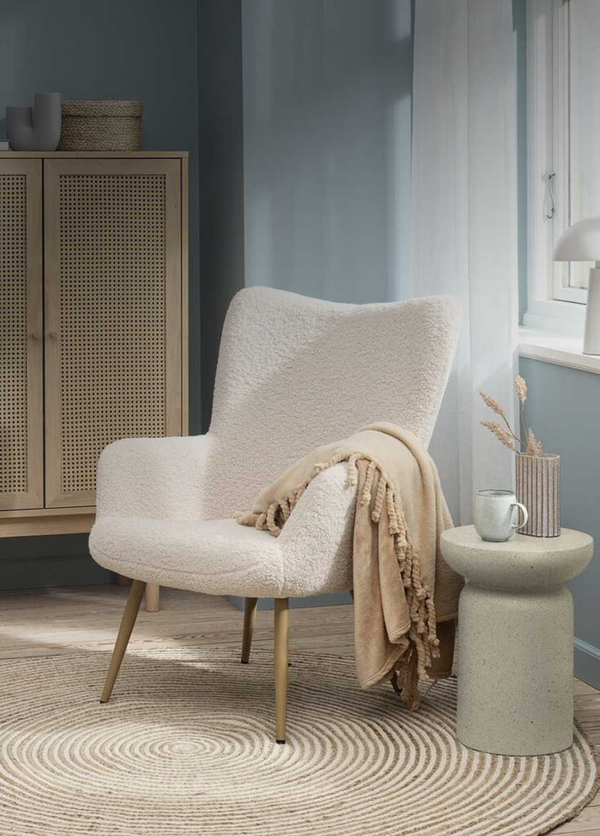Armchairs Cream bouclé wingback armchair with gold metal legs and a beige throw, beside a rattan wardrobe and a stone side table.