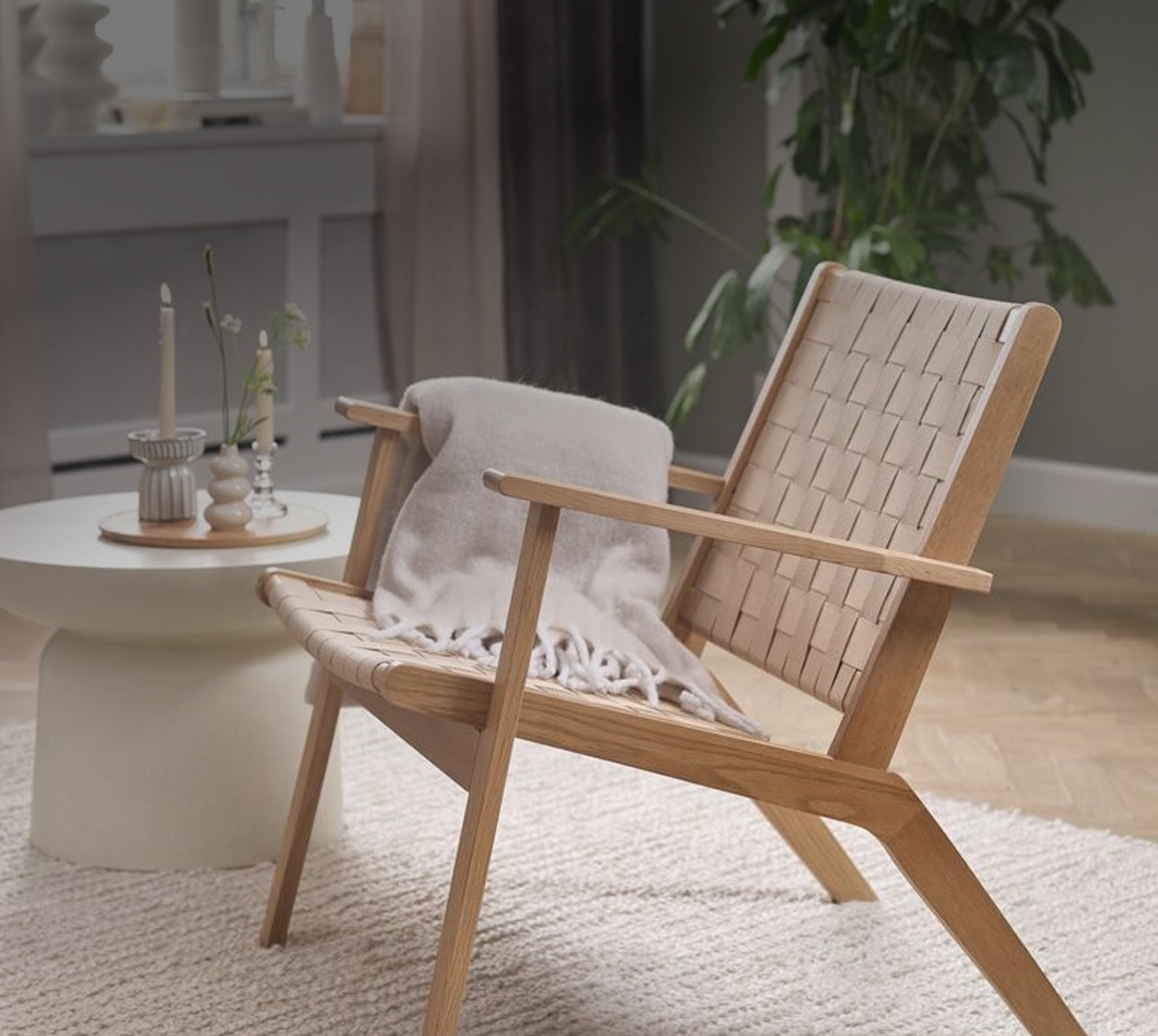 Armchairs Oak-framed armchair with a woven beige strap seat and backrest, draped with a cream throw, beside a round side table with candles and flowers in a living room.