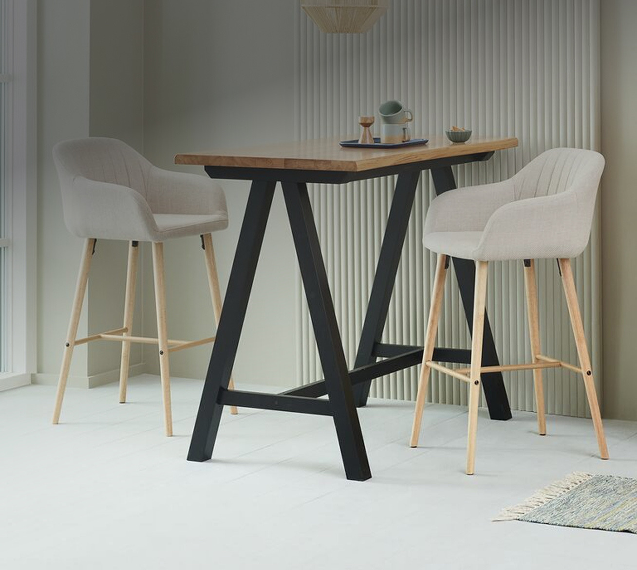 Bar stools and tables Two beige upholstered bar stools with wooden legs at a black A-frame bar table set with mugs and a tray, in front of a ribbed wall panel.