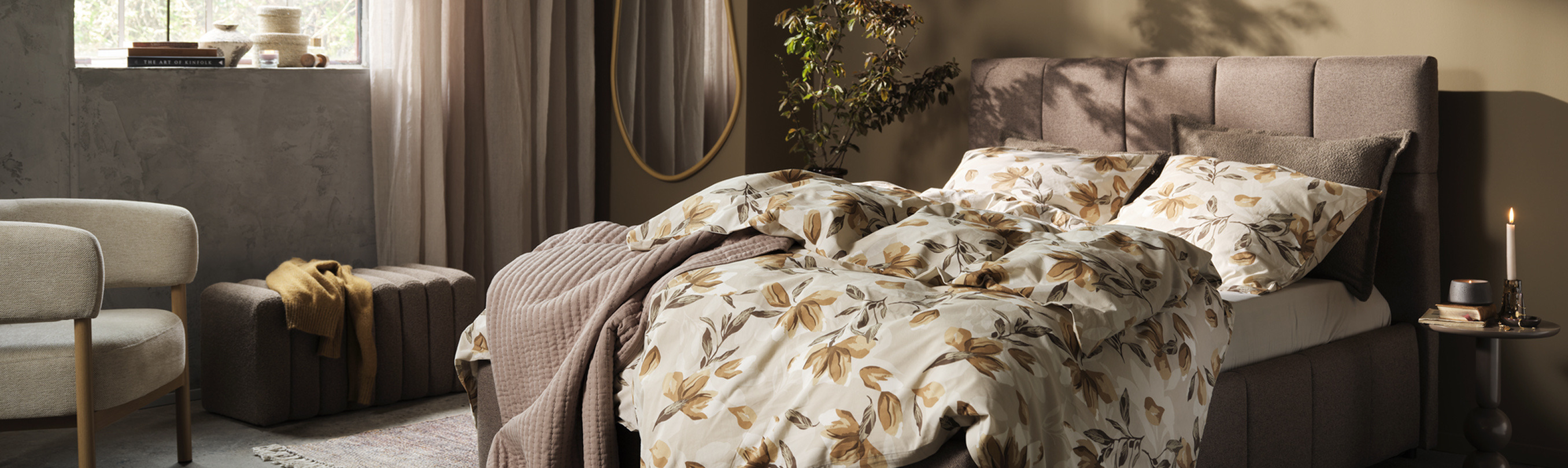 Bedroom shopping guide cover Bedroom with a taupe upholstered bed dressed in a cream and rust floral duvet, a beige armchair, a round mirror, and a potted plant, lit by warm ambient light.