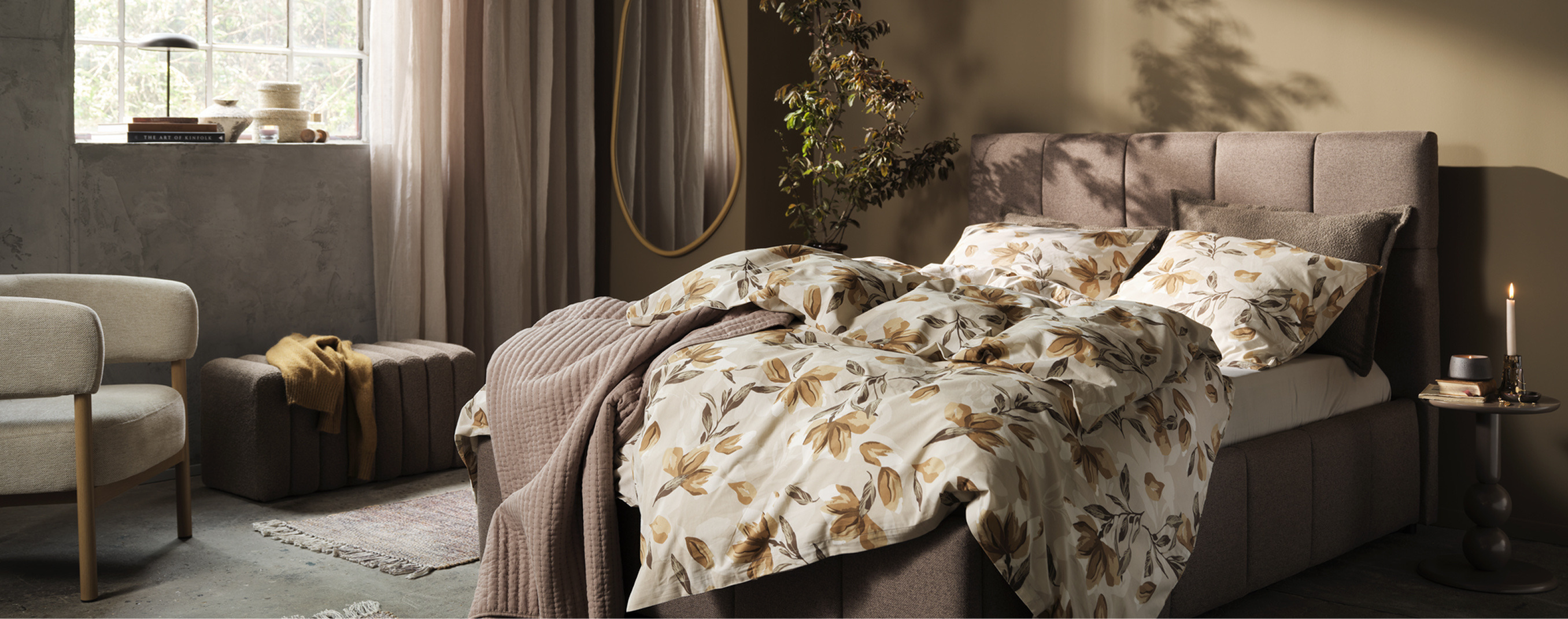 Bedroom shopping guide mobile cover Bedroom with a taupe upholstered bed dressed in a cream and rust floral duvet, a beige armchair, a round mirror, and a potted plant, lit by warm ambient light.