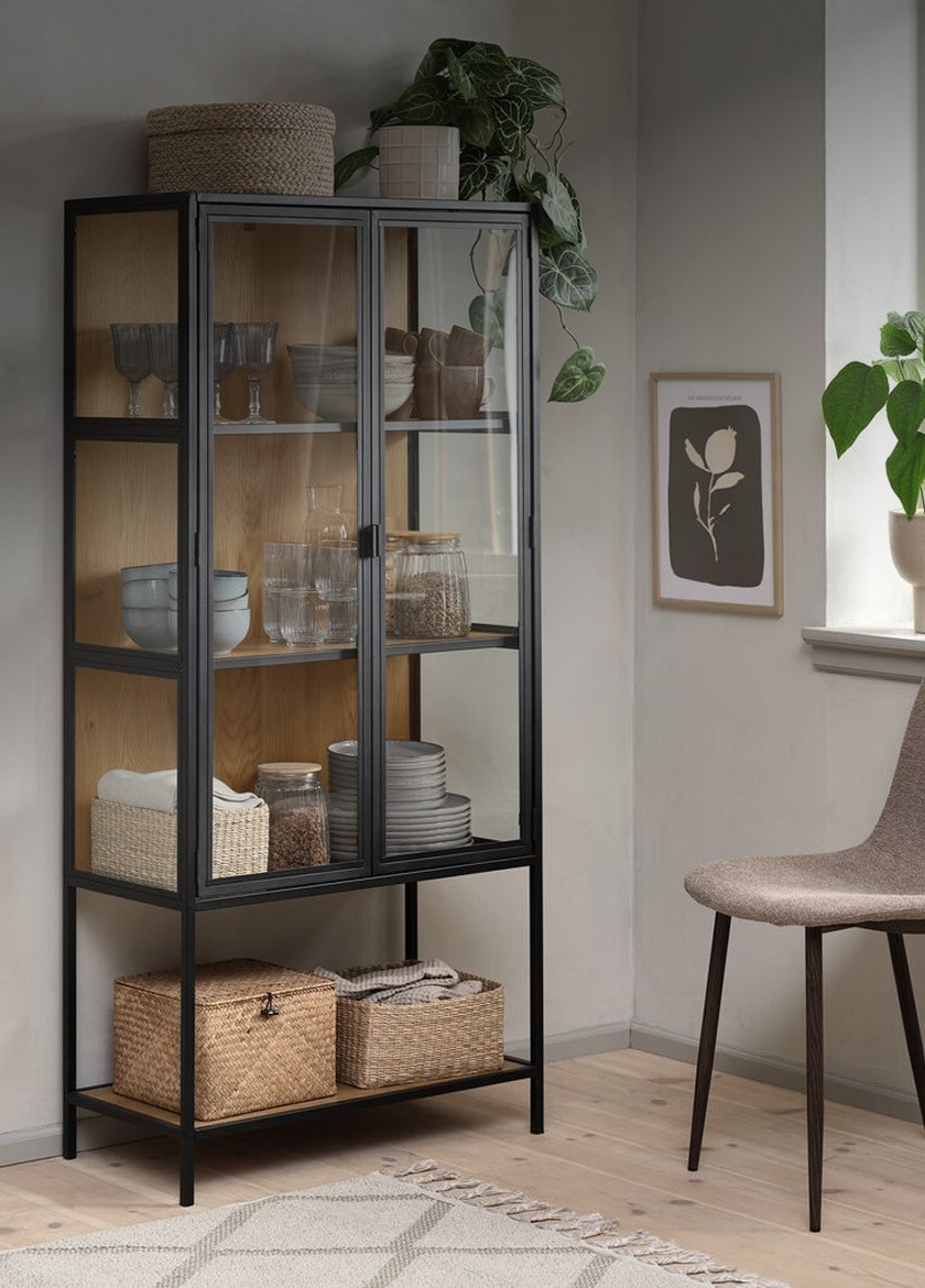 Cabinets Black-framed glass display cabinet with wood-backed shelves holding glassware, bowls, and jars, beside a potted plant and framed art.