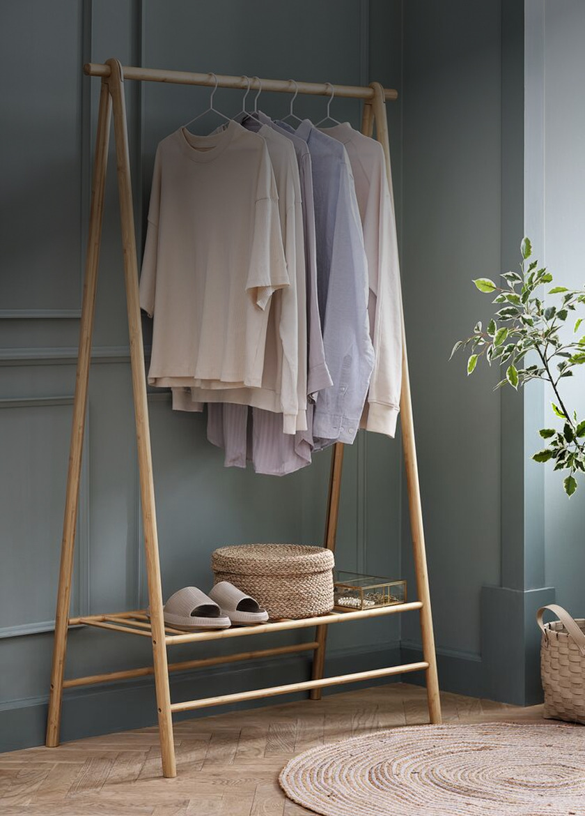 Clothes rails Freestanding wooden A-frame clothes rail with folded sweaters and shirts on hangers, slippers and a woven basket on the lower shelf, beside a potted plant.