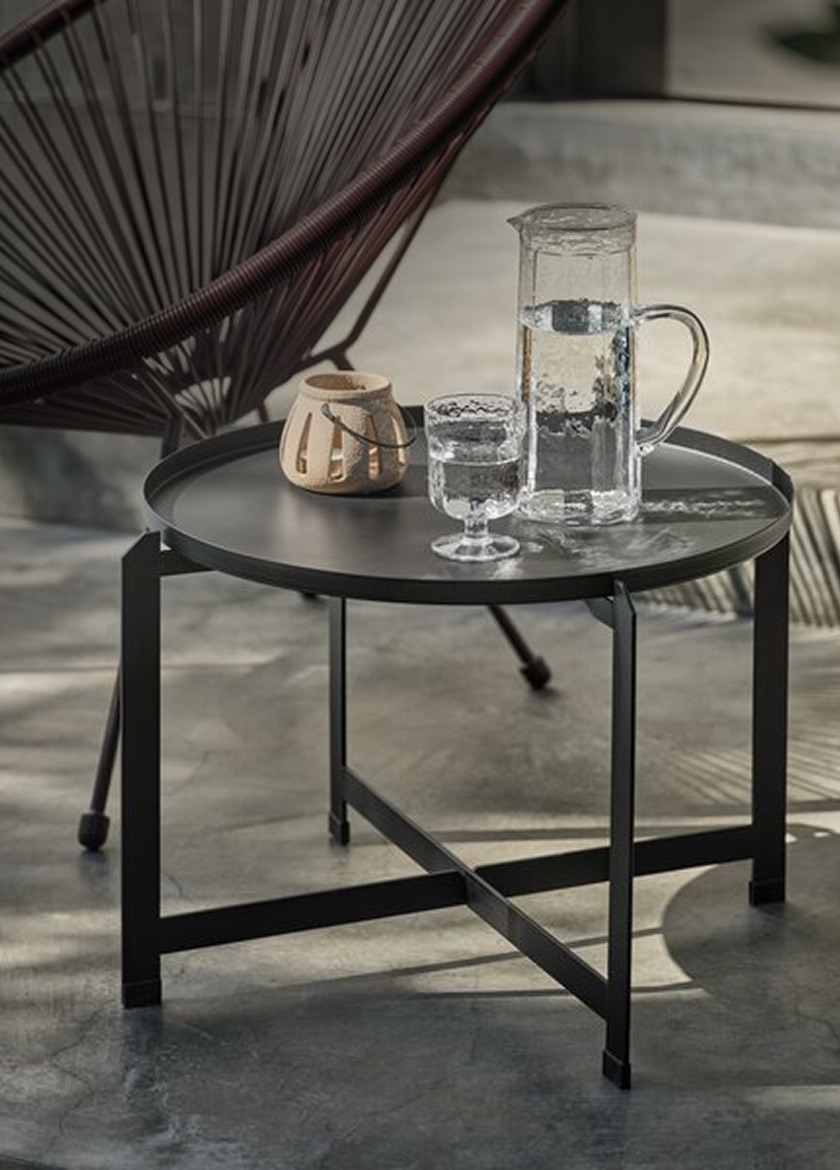 Coffee tables Round black metal coffee table with a cross-brace base, set with a glass jug of water, a glass, and a small vase, beside a woven egg chair.