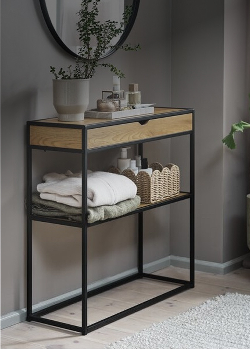 Console tables Round grey three-legged side table with a ribbed glass vase of dried eucalyptus stems, in front of a wood-paneled wall.