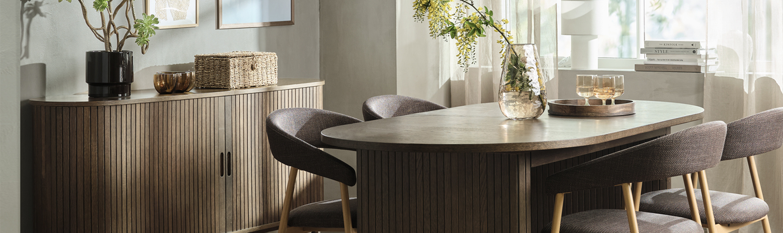 Dining room cover Dining room with a ribbed wood sideboard, framed art, and an oval wood table surrounded by upholstered chairs, with a glass vase of flowering branches.