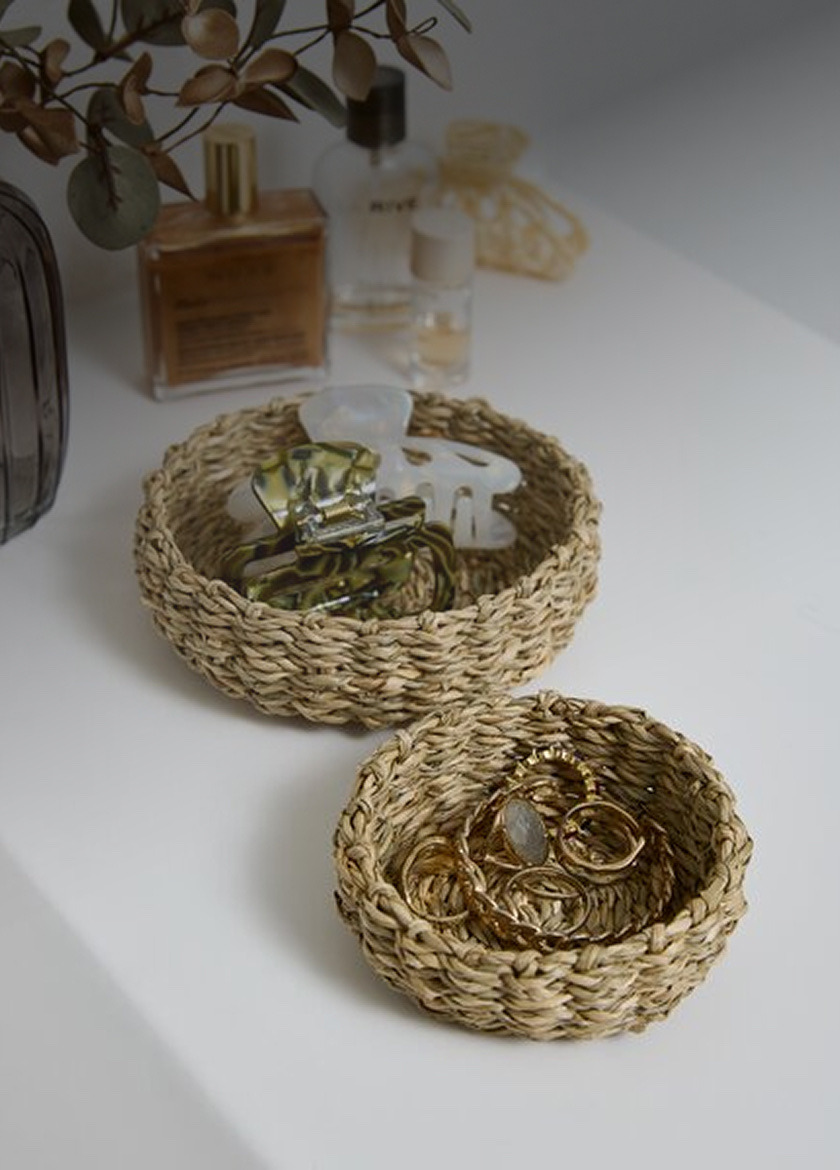 Extra accessories Two woven seagrass trinket bowls holding hair clips and gold jewelry, beside perfume bottles on a white shelf.