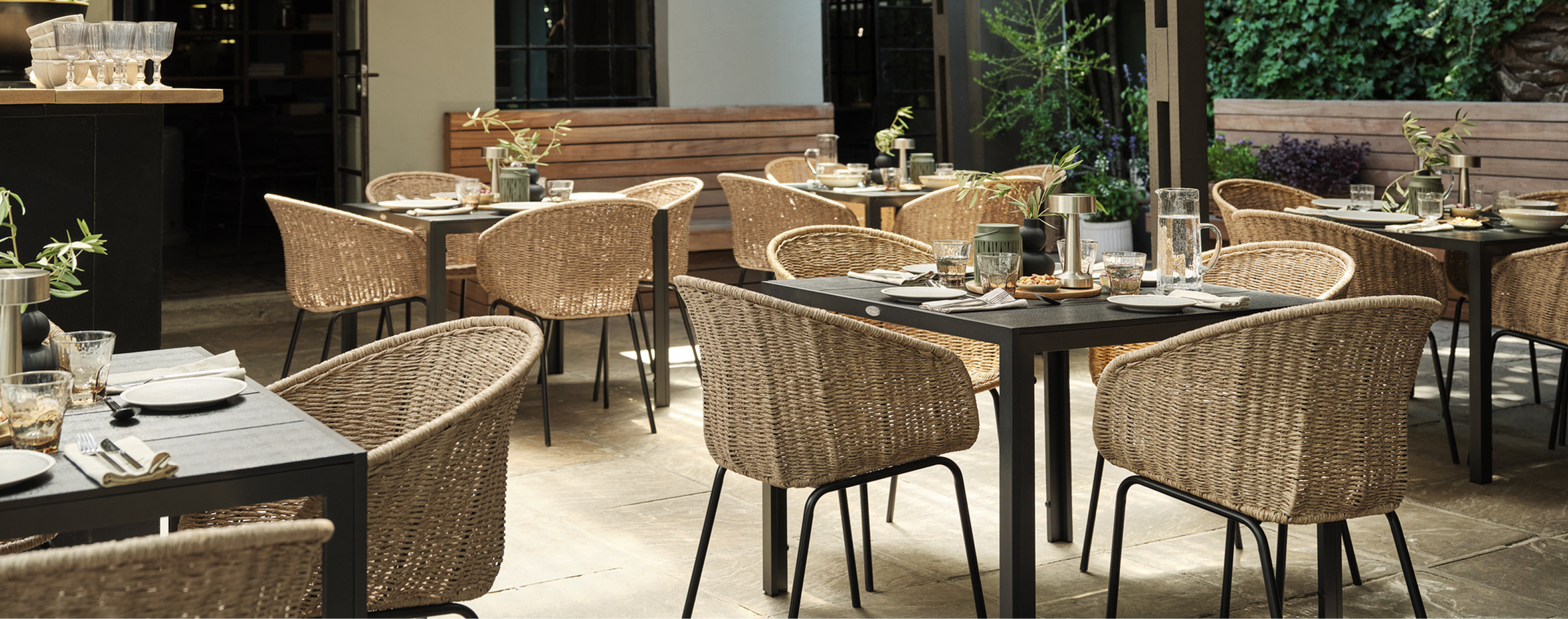 Garden cover Outdoor garden dining terrace with wicker chairs around dark tables set for a meal, surrounded by greenery and warm lighting.