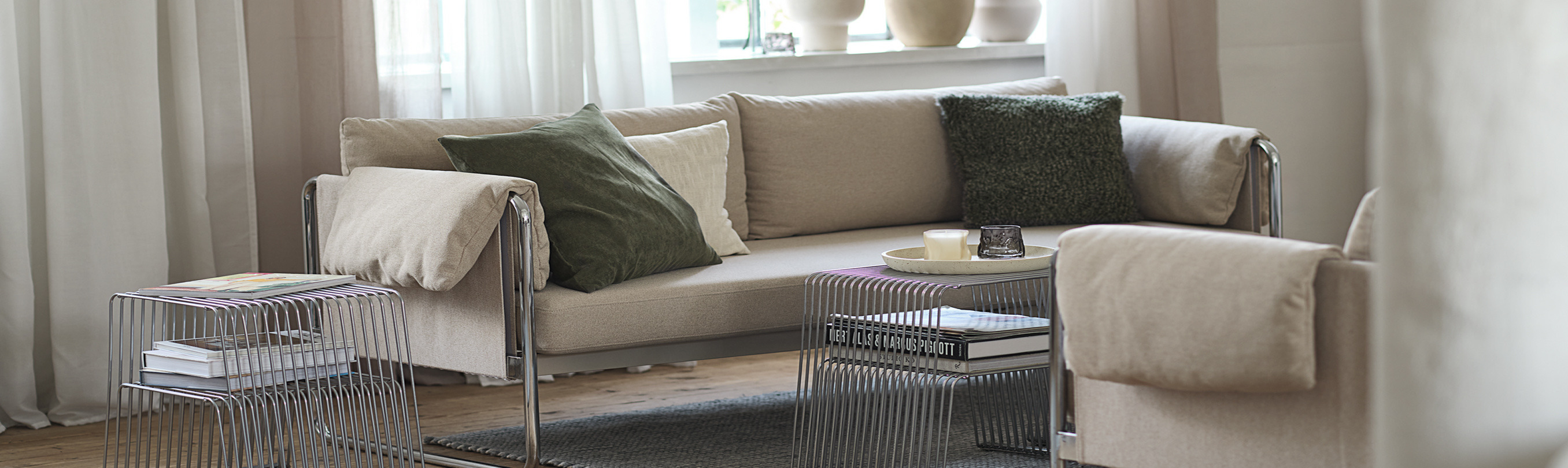 Living room cover Living room with a beige sofa and armchair, green and cream cushions, sheer curtains, and two wire-frame side tables with magazines.