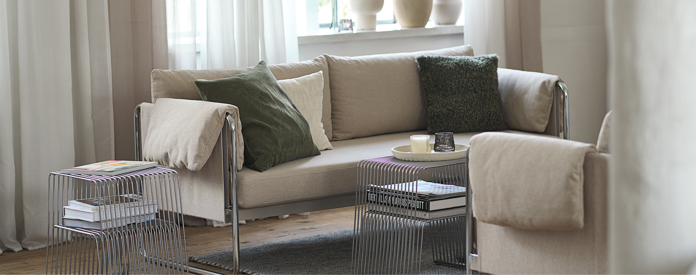 Living room cover Living room with a beige sofa and armchair, green and cream cushions, sheer curtains, and two wire-frame side tables with magazines.
