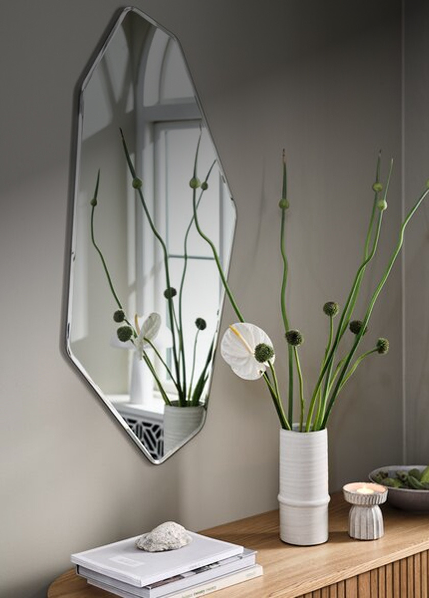 Mirrors Angular diamond-shaped wall mirror above a wood shelf styled with a vase of greenery, a candle, and books.