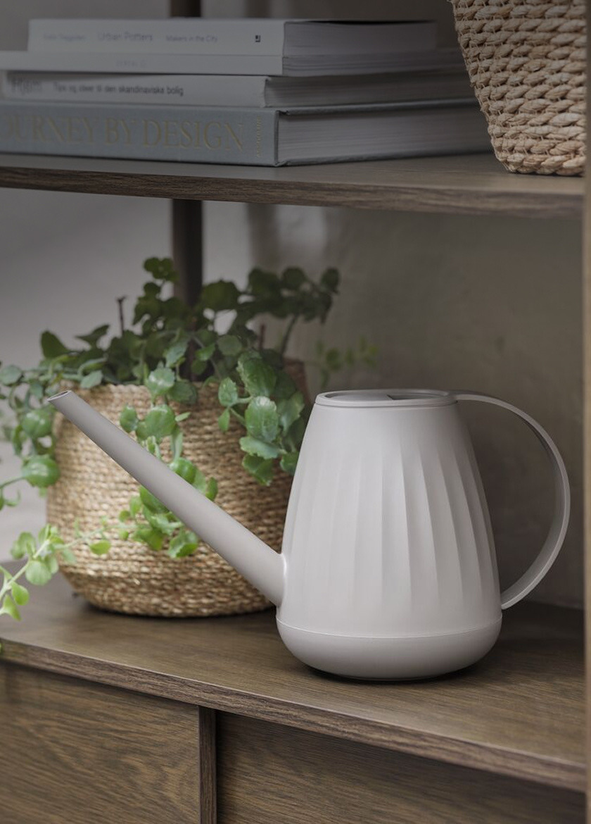 Miscellaneous Grey watering can with a long spout, beside a woven basket planter and a stack of books on a wood shelf.
