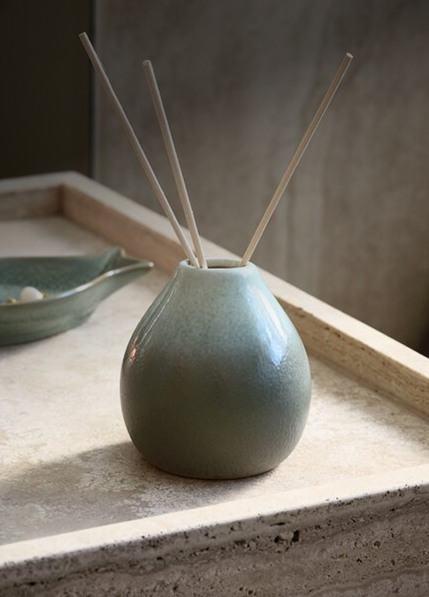 Reed diffusers Teal ceramic reed diffuser with wooden reed sticks, on a stone tray beside a small dish, in soft natural light.