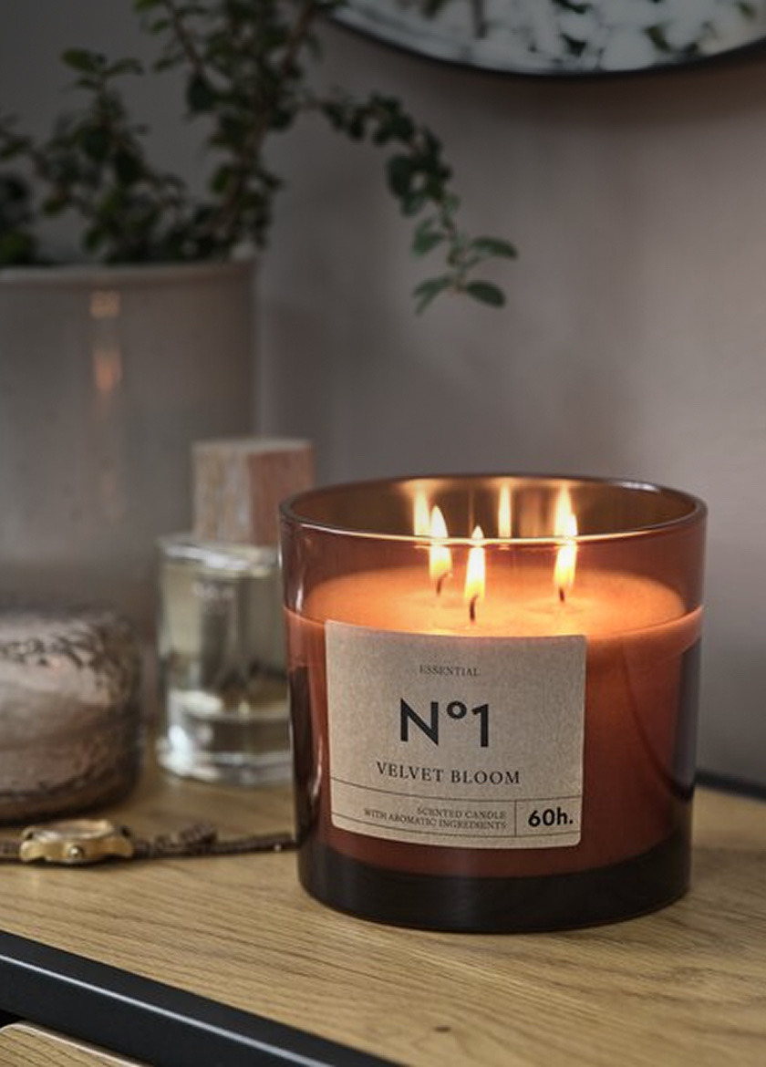 Scented candles Amber glass jar candle labeled 'No1 Velvet Bloom' with three lit wicks, on a wood shelf beside a diffuser bottle and a potted plant.