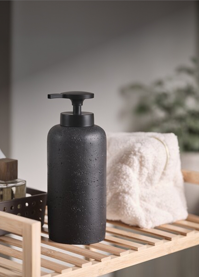 Soap dispensers Dark textured stone soap dispenser bottle beside a folded white towel and a potted plant on a wood shelf.