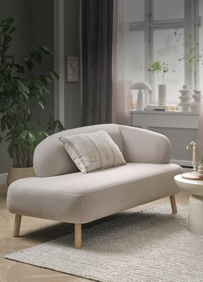Sofas Beige upholstered two-seater sofa with a curved backrest and a striped accent pillow, in a living room with plants and a round side table.