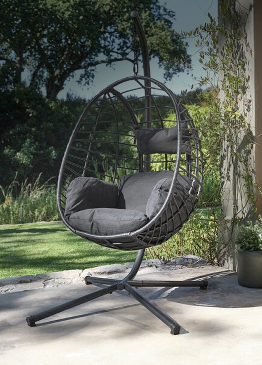 Swing chairs Black wicker hanging egg chair with grey cushions on a curved metal stand, on an outdoor patio beside a potted plant.