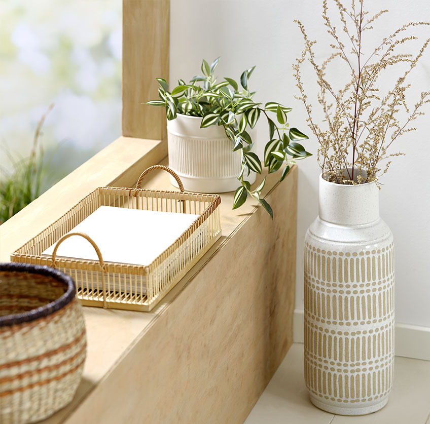 Große Vase neben einer Fensterbank, darauf ein Tablett aus Bambus und weisser Pflanzentopf mit Kunstpflanze darin