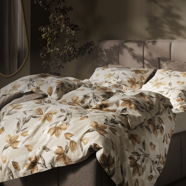 Bedroom with a taupe upholstered bed dressed in a cream and rust floral duvet, a beige armchair, a round mirror, and a potted plant, lit by warm ambient light.