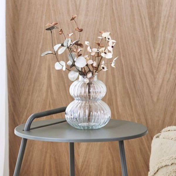 Artificial flowers Clear ribbed glass vase with artificial dried eucalyptus and flower stems, on a grey round side table against a wood-paneled wall.
