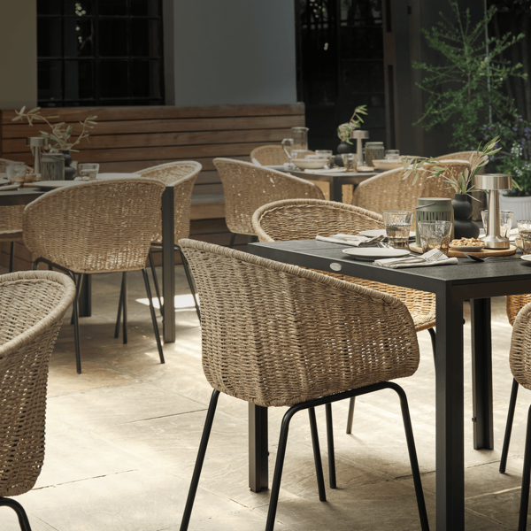 Outdoor garden dining terrace with wicker chairs around dark tables set for a meal, surrounded by greenery and warm lighting.