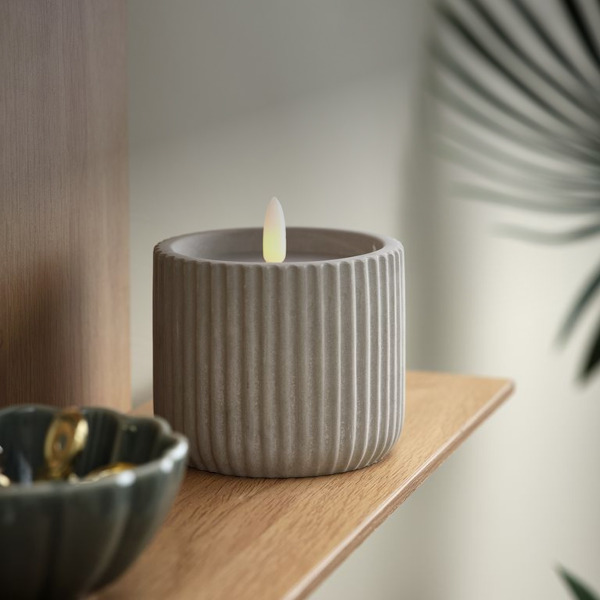 LED candles Grey ribbed ceramic pot holding a flameless LED candle, on a wood shelf beside a dark bowl with gold jewelry.