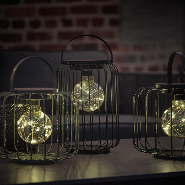 Lamps and lanterns Three black wire cage lanterns of varying heights holding glowing string-light bulbs, on an outdoor table at dusk.