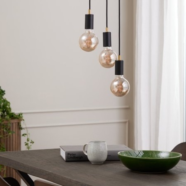 Lamps and light bulbs Three black pendant lights with exposed round filament bulbs hanging above a dark wood dining table set with a book, a mug, and a green ceramic bowl.