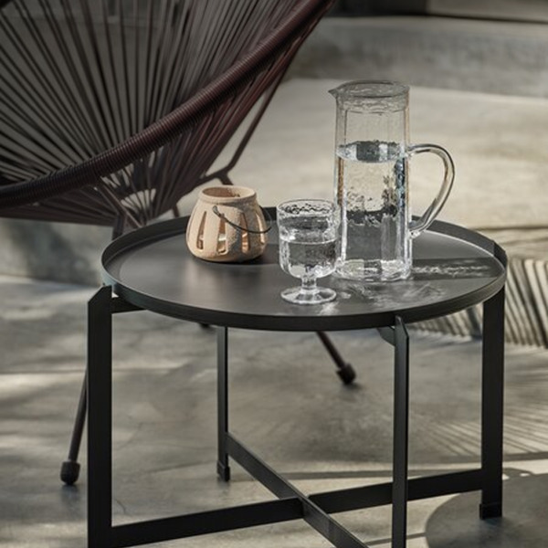 Lounge tables Round black metal tray-top side table set with a glass water pitcher, a glass, and a small ceramic lantern, beside a woven egg chair.