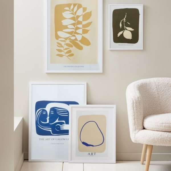 Picture frames White picture frames leaning against a wall and hanging above, holding botanical and abstract art prints in yellow, blue, and olive tones, beside a cream bouclé armchair.