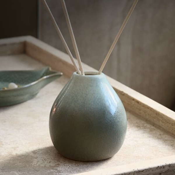 Reed diffusers Teal ceramic reed diffuser with wooden reed sticks, on a stone tray beside a small dish, in soft natural light.