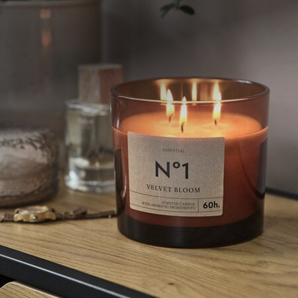 Scented candles Amber glass jar candle labeled 'No1 Velvet Bloom' with three lit wicks, on a wood table beside a potted plant.