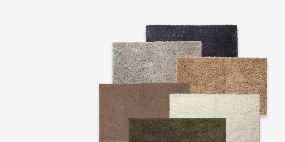 Bath mats Stack of layered bath mats in black, grey, taupe, beige, cream, and olive green, shown against a plain light background.