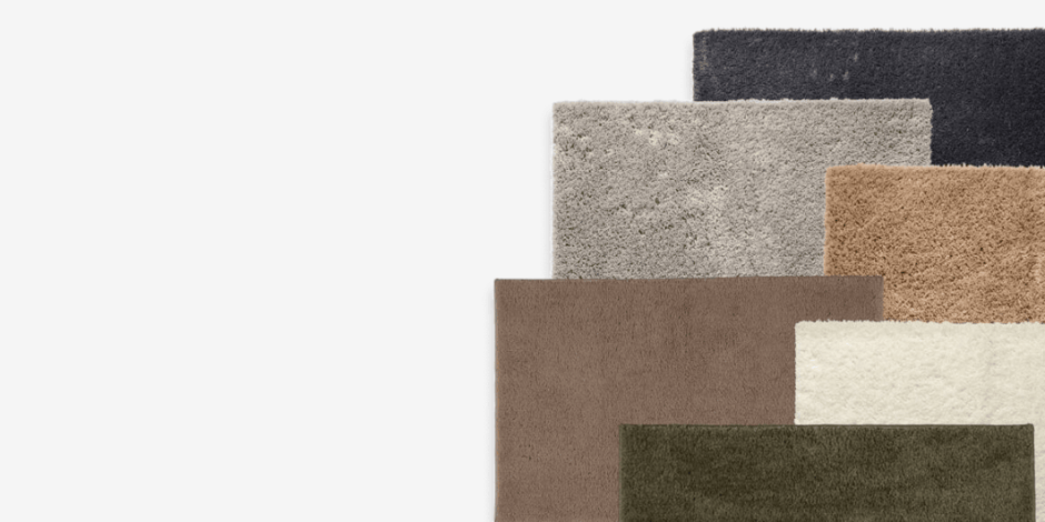 Bath mats Stack of layered bath mats in black, grey, taupe, beige, cream, and olive green, shown against a plain light background.