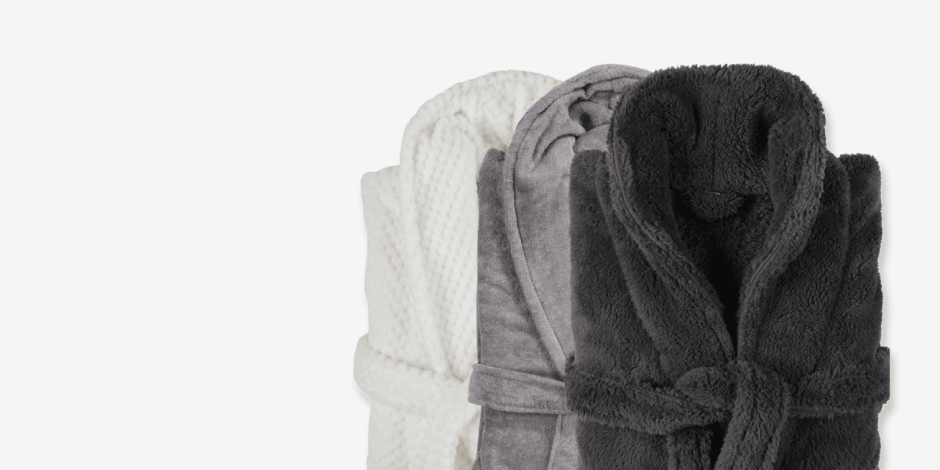 Bathrobes Three rolled bathrobes in white, grey, and charcoal plush fabric, shown against a plain light background.