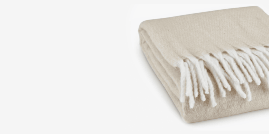Throws Folded beige knit throw blanket with a fringed edge, shown against a plain white background.