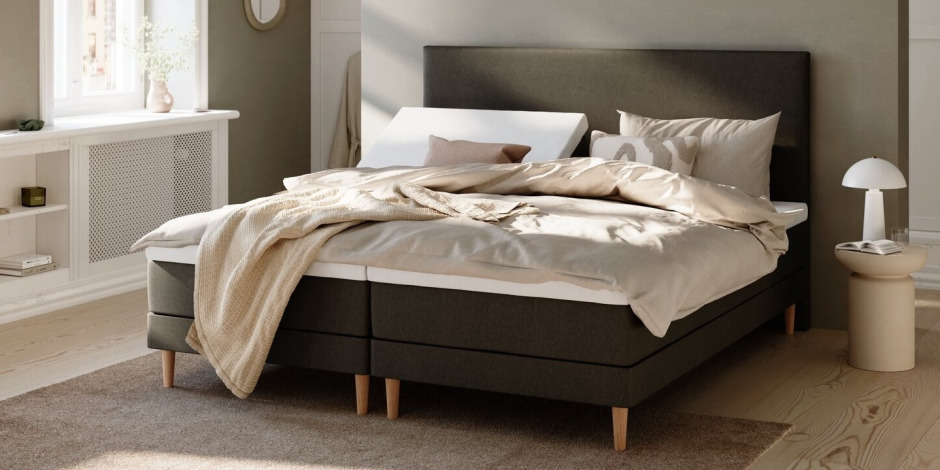 Complete bed sets Dark grey upholstered bed with beige linens, a folded knit throw, striped accent pillows, and a round bedside table with a mushroom lamp in a sunlit bedroom